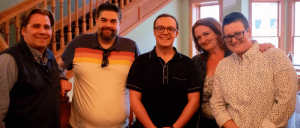 Group of five people posing indoors with a wooden staircase in the background.