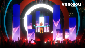 A vibrant virtual concert stage setup featuring a DJ booth with a glowing holographic performer at center stage, surrounded by tall illuminated blue rectangular pillars and a large white circular arch backdrop. The foreground shows silhouettes of a cheeri