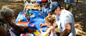 A group of people engaged in a craft activity with paper and markers on a blue mat outdoors.