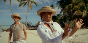 Still photo from Waltzing with Brando featuring Billy Zane as Marlon Brando and Jon Heder. Zane, in a white shirt, scarf, and straw hat, gestures on a tropical beach. Heder, in a striped shirt and hat, stands behind him, looking uncertain amid palm trees."