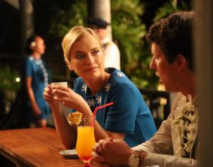 Still photo from Waltzing with Brando with Camille Razat as Michelle and Jon Heder as Bernard Judge. Razat, in a blue floral dress, holds a cup while listening to Heder, in a light suit with a lei. They sit at an outdoor bar with tropical decor and warm lighting.