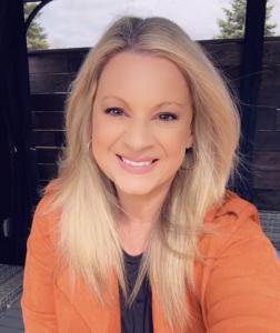 A professional photo of Katharine Loucaidou, a blonde woman wearing an orange blazer and smiling confidently, set against a natural outdoor backdrop. She is a globally recognized AI strategist and business coach.