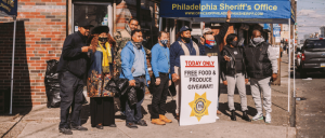 A group of people stands in front of a tent witA community safety officer engages with a citizen amidst a bustling urban scene with police officers and pedestrians in the background a sign announcing a free food and produce giveaway event.