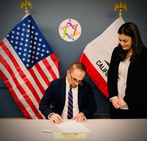 A man in a suit is sitting at a table, signing a document, with a woman standing beside him. Behind them are the U.S. and California flags and a colorful circular logo on the wall.