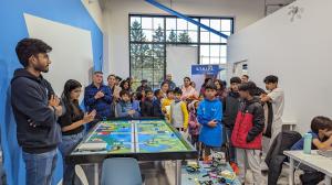 A group of students, parents, and mentors stand around a robotics challenge table at the STRIPE Competition. Some are clapping, while others observe the event with excitement. The competition banner is visible in the background, along with various LEGO ro