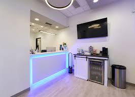 Reception area at Love Dental with a sleek front desk illuminated by blue lighting, a refreshment station with a beverage fridge, and a mounted TV for patient comfort.