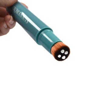 A close up of a sunscreen applicator in the colour Sea Jewel with white sunscreen being pumped from the bristles is held by a person, illustrating the use of the SUNNYBOD refillable sunscreen brush applicator.