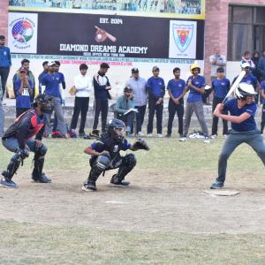 A baseball practise game in progress at Diamond Dreams Academy. The batter is ready to swing, with the catcher and umpire crouched behind home plate. Several spectators and participants stand in the background near a large banner