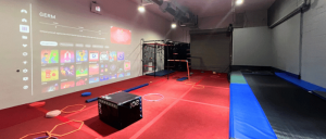 A brightly lit gym with a projected screen displaying colorful graphics, red flooring, and various fitness equipment scattered throughout.