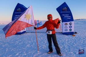 "A woman in a red jacket, black pants, and sunglasses stands on a snowy landscape, holding the Philippine flag with one hand and making a peace sign with the other. Behind her, blue banners mark the start of the World Marathon Challenge in Antarctica, wit