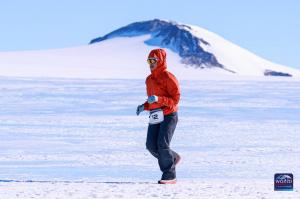 A woman in a red jacket, black pants, and sunglasses runs across a vast, icy landscape in Antarctica. She wears race bib number 12, with a snow-covered mountain in the background. The bright sunlight casts long shadows on the snow as she competes in the W