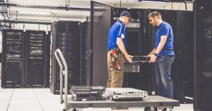 Two OceanTech technicians in blue shirts are carefully removing a server from a rack. One technician is wearing a cap and tool belt, while the other is wearing sunglasses and a smartwatch. A rolling cart nearby holds additional server equipment. The backg