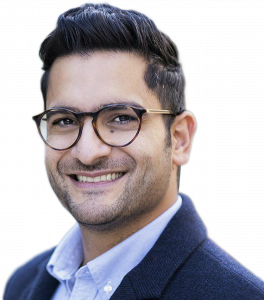 Professional headshot of a smiling man wearing round tortoiseshell glasses, a light blue button-down shirt, and a navy sweater or blazer. He has dark hair styled neatly and maintains a well-groomed beard."