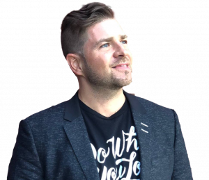 "Professional headshot of a man looking up and to the side with a slight smile. He has short brown hair, light stubble, and is wearing a textured navy blazer over a black t-shirt with white text. The image is cropped at chest level and set against a white