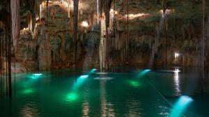 A breathtaking underground cenote with crystal-clear turquoise waters, surrounded by stalactites and stalagmites. Sunlight filters through the cave ceiling, illuminating the rock formations and a spiral staircase leading into the depths.