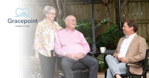 An elderly man in a wheelchair, a smiling elderly woman, and a younger man in a blazer sit in a lush outdoor garden, enjoying a friendly conversation. The Gracepoint Home Care logo is visible on the left side.