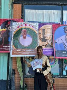 Tami Outterbridge, daughter of the late John Outterbridge, stands in front of a tribute banner honoring her father at the 14th Annual Day of the Ancestors: Festival of Masks in Leimert Park. The festival celebrated the legacy of cultural icons like John O