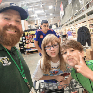 A group of children smiles at the camera while shopping in a grocery store, with shelves of products in the background.