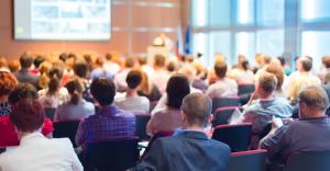 A large audience is seated in a conference hall, attentively listening to a speaker at the front of the room. The speaker stands behind a podium, delivering a presentation with a projected screen displaying images. The attendees, dressed in business casua