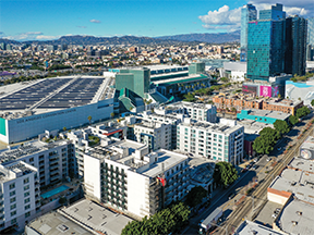 An aerial view of Liv DTLA, a modern mid-rise residential building in Downtown Los Angeles. The building is surrounded by other contemporary apartment complexes, the Los Angeles Convention Center, and city streets lined with trees. In the background, the