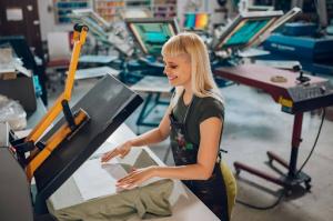 A blonde woman screen printing a shirt in a workshop.