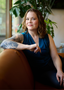 Sara Lobkovich is sitting sideways on a brown sofa. Behind her is greenery and a window. She's looking in slight profile at the camera with a casual smile on her face. She has blue eyes and wavy dark blonde hair. She's wearing a teal v-neck sleeveless top