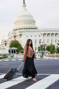 A woman with long brown hair walks across a crosswalk in front of the U.S. Capitol, pulling a black suitcase. She wears a black dress, a brown jacket, and gold high heels, looking toward the camera with a confident expression. The background features the 