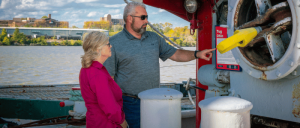 A man points at a control panel on a tugboat while a woman in a pink shirt listens, with a river and trees in the background.