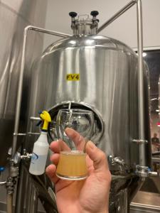 A hand holds a small glass filled with a hazy IPA, sampled directly from a large stainless steel fermentation tank labeled "FV4" during the brewing process. The tank, part of Westminster Brewing Company's setup, is surrounded by industrial brewing equipme