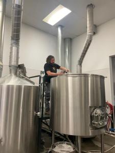 A photo of Ryan, a brewer at Westminster Brewing Company, standing on a platform as he works on his first hazy IPA. He is positioned in front of a large stainless steel fermentation tank, surrounded by industrial brewing equipment, including pipes and add