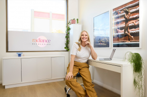 Ally Dellabarca, founder of RadianceRevolution.com.au, sitting and smiling happily with the Radiance Revolution background, reflecting her passion for beauty and wellness innovation.