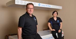 David Linton, NP, sits on the left while Kezia Badulid sits on the right, both wearing Long Life Med-branded uniforms in their clinic. They are sitting on top of the fully body NIR and Red Light Therapy Bed, with another Long Life Med RLT panel above them