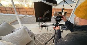 Photographer Matt Janson, wearing a mustard yellow beanie, adjusts a camera mounted on a slider while capturing a 3D image of a frost-covered flower. The setup includes a black backdrop, studio lighting, and a tripod, all arranged on a covered patio with 