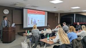 Dr. Kurusu speaks at a podium while a group of attendees listens to a lecture on elderly care, shown on a projector screen.