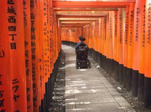 Josh Grisdale, in his power wheelchair, moves through the torii gate tunnel at Fushimi Inari Shrine in Kyoto, Japan. The bright orange gates line the stone pathway.