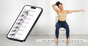 A woman with curly hair sits on a chair performing a seated side neck stretch, wearing a beige tank top and navy leggings. She is on a STRETCHIT-branded mat in a bright, minimalist space. To the right, a large floating smartphone screen displays the STRET