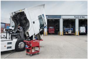 A diesel mechanic performing maintenance on a truck in front of a professional, well-established truck repair shop. The setting includes a clean, organized workspace with visible tools and signage, representing high-quality service and expertise.