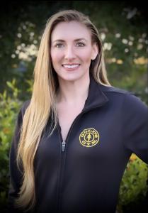 Tiffany stands in a black Gold's Gym jacket and smiles outdoors with greenery in the background.