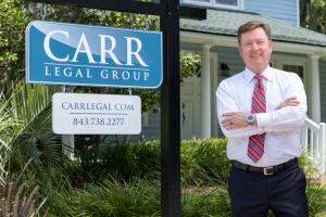 Patrick W. Carr, a professionally dressed man with short brown hair, wearing a white dress shirt, red and blue striped tie, and a wristwatch, stands confidently with his arms crossed in front of a blue and white sign that reads 'CARR LEGAL GROUP.' The sig