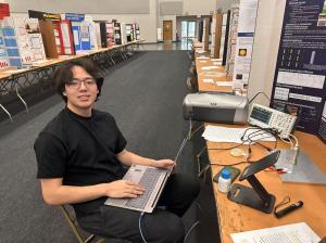 Max Kopp, a 17-year-old scientist, sits at a science fair working on his AI-powered nanotechnology project for spacecraft safety. His setup includes a laptop, scientific instruments, and research posters.