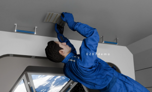 An astronaut in a blue spacesuit installing a flexible nanomaterial sensor strip on the interior surface of a spacecraft. The Earth is visible through a window, and the technology is designed for real-time structural health monitoring.