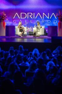 Two women seated on stage in white chairs, engaged in a conversation with microphones in hand. One wears a black dress, the other a shiny silver suit. A large audience watches attentively below in the foreground.