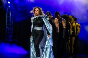 A woman in a black bodysuit with a silver metallic cape walks confidently across a fog-filled stage, leading a line of dancers dressed in black. Blue and purple lights create a dramatic, theatrical atmosphere.