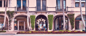 Two shoppers walk under archways of a stylish Mediterranean-style building, featuring lush greenery and elegant storefronts.