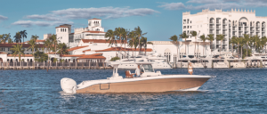 A sleek boat glides through calm waters beside a tropical waterfront, featuring elegant buildings and palm trees under a clear sky.