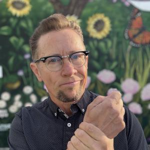 White older guy with light brown hair and browline glasses in short sleeved shirt standing in front of a flower mural with one hand around his other wrist.