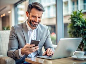 man in business attire smiling at laptop in coffee shop while holding phone