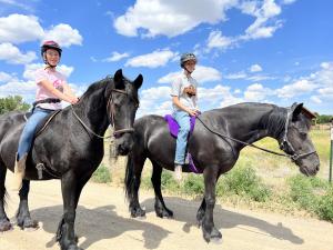 Two students smiling while sitting on horses at the ranch, enjoying their lesson and bonding with the animals in a supportive, outdoor environment.