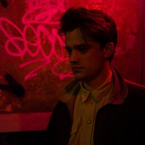 A young man stands in the glow of a red neon haze. There is some graffiti on the wall behind him which isn't legible. The man is stood in a slight side profile, looking to the left of the image.