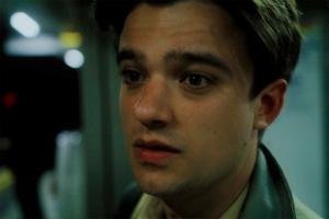 A close-up image of a young man who appears slightly emotional, as if pleading or begging. The man is actor Adam Wadsworth, who portrays Victor in the film Nosebleed. The image has cold tones, with a bluey-green tint.